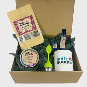 May include: A gift box filled with self-care items. Includes a tea bag, a mug with the text "holly botanic", a tea infuser, a balm tin, and essential oil bottles. The box is lined with green tissue paper.