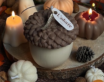 May include: A collection of autumn-themed candles, including an acorn, pumpkins, and a ghost. The acorn candle is brown and cream, with a tag that reads "L'adorée". Other candles are orange, white, and brown. The scene is decorated with fall leaves and fairy lights.