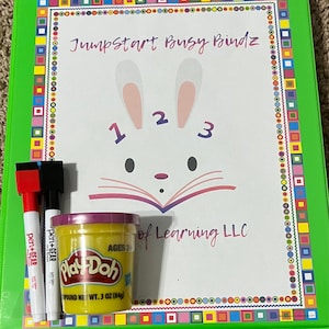 May include: A green binder with a colorful border and a white page with a bunny illustration and the text "Jumpstart Busy Bindz of Learning LLC". The page also has the numbers 1, 2, and 3. A container of Play-Doh is in the foreground.