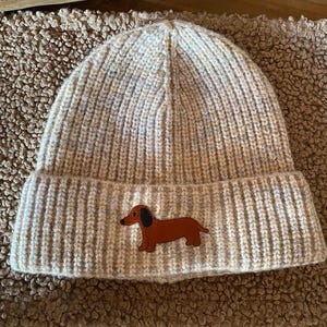 Könnte beinhalten: Eine beigefarbene, gerippte Strickmütze mit einem kleinen aufgestickten Aufnäher eines braunen Dackels mit schwarzem Ohr. Die Mütze verfügt über einen breiten Umschlag und eine texturierte, gesprenkelte Optik im gesamten Strickgewebe.