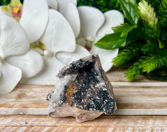 May include: A raw, uncut crystal geode with a dark interior speckled with white and yellow accents. The geode sits on a wooden surface, with white orchid flowers and green foliage in the background.