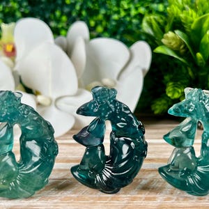 May include: Three teal blue carved crystal fish sculptures with intricate details. The fish are arranged in a semi-circle on a wooden surface.