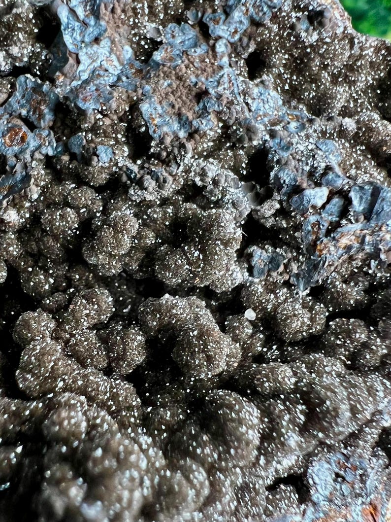 Op de afbeelding: Close-up van een donkergrijs en zilverkleurig mineraalmonster met een ruw, getextureerd oppervlak. Het mineraal heeft een glinsterende, kristallijne uitstraling.