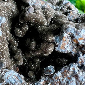 Op de afbeelding: Close-up van een donkergrijze, getextureerde steen met een zilveren, iriserende glans. De steen heeft een ruw, onregelmatig oppervlak met veel kleine, onregelmatige vormen.