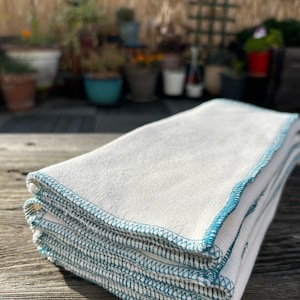 Peut inclure: Une pile de torchons en coton blanc avec des coutures bleu turquoise autour des bords. Les torchons sont pliés et empilés soigneusement.