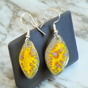 May include: A pair of dangle earrings with silver hooks and yellow and purple abstract designs. The earrings are displayed on a gray background.