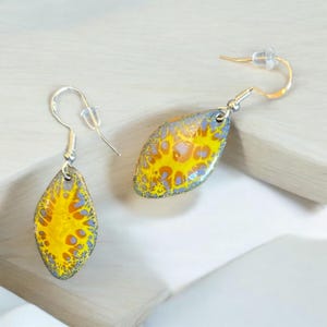 May include: A pair of silver dangle earrings with yellow and blue abstract designs. The earrings are shaped like a teardrop.