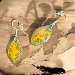 May include: A pair of silver dangle earrings with yellow and blue abstract designs. The earrings are shaped like teardrops.