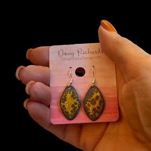 May include: A pair of yellow and black enamel earrings with a silver hook. The earrings are shaped like a teardrop and have a marbled pattern. The earrings are displayed on a pink card with the text "Doug Richards OriginalsbyDoug.com".
