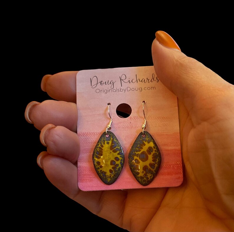 May include: A pair of yellow and black enamel earrings with a silver hook. The earrings are shaped like a teardrop and have a marbled pattern. The earrings are displayed on a pink card with the text "Doug Richards OriginalsbyDoug.com".