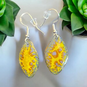May include: A pair of silver dangle earrings with yellow and gray abstract designs. The earrings are teardrop shaped and have a silver hook.