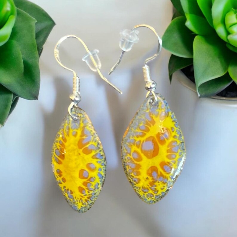May include: A pair of silver dangle earrings with yellow and gray abstract designs. The earrings are teardrop shaped and have a silver hook.
