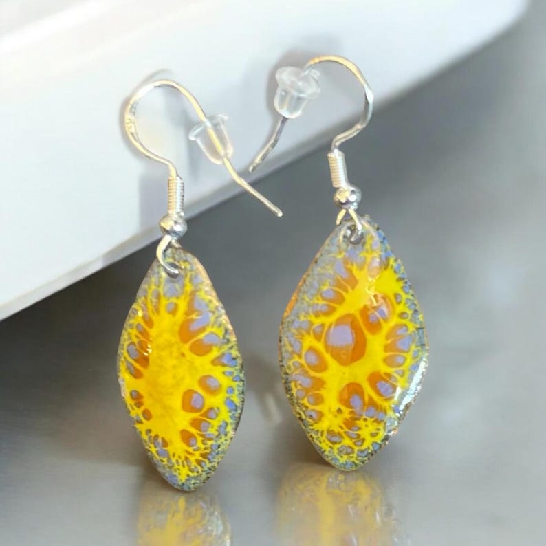 May include: A pair of silver dangle earrings with yellow and purple enamel teardrop-shaped pendants. The pendants have a marbled, abstract design.