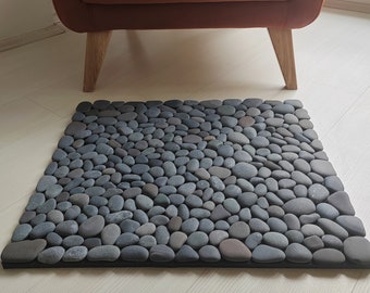 May include: A gray stone bath mat with smooth, rounded stones arranged in a close pattern. The mat is placed on a light wood floor in front of a brown armchair.