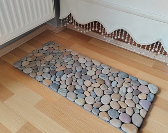 May include: A rectangular bath mat made of natural stones in various shades of brown, gray, and beige. The stones are arranged in a random pattern, creating a textured surface.