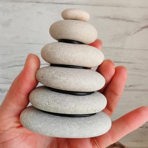 May include: A stack of six smooth, light gray stones, each oval-shaped, balanced on top of each other. Black rings separate the stones. The top stone is a lighter color. The stones are held in a hand.