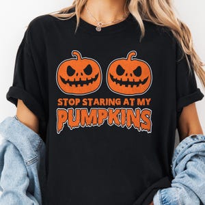 May include: Black t-shirt featuring two orange jack-o'-lanterns with carved faces. The text "STOP STARING AT MY PUMPKINS" is printed in orange below the pumpkins. A Halloween-themed graphic tee.