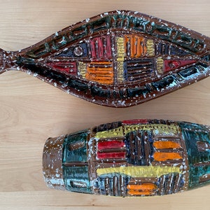 May include: A set of two ceramic vases with a brown, green, yellow, orange, and red geometric pattern. The vases have a speckled glaze and are in the shape of a fish and a cylinder.