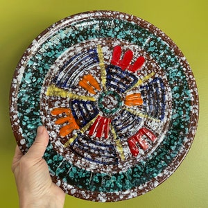 May include: A round ceramic plate with a colorful geometric design. The plate has a brown background with white speckles and a central design of blue, red, orange, and yellow lines and shapes. The outer edge of the plate has a green and white speckled design.