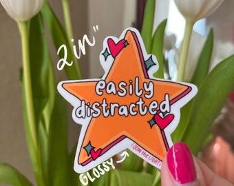 May include: A 2 inch glossy sticker with an orange star shape. The star has a white outline and the text "easily distracted" in black. The sticker has a pink heart and a blue star on each point. The sticker has the text "glow the light" in white on the bottom.