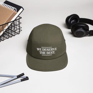 May include: An olive green five-panel cap with a flat brim. The cap features the white text "WE DESERVE THE BEST." Black headphones and a wire basket with notebooks are in the background.