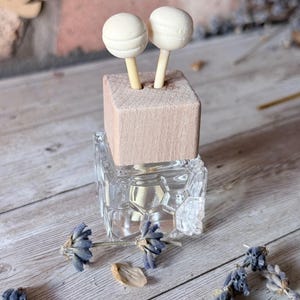 May include: A clear glass diffuser bottle with a light brown wooden top. Two white, spherical diffuser sticks are inserted into the wooden top. Dried lavender sprigs and petals are scattered around the bottle on a wooden surface.