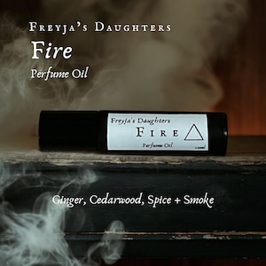 May include: A black roller bottle with a white label that reads "Freyja's Daughters FIRE Perfume Oil 10ml". The bottle is sitting on a dark wooden surface with white smoke in the background. The text "Ginger, Cedarwood, Spice + Smoke" is written below the bottle.