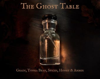 May include: A clear glass perfume bottle with a black cap, labeled "The Ghost Table." The bottle contains a light-colored liquid and a spray mechanism. The bottom of the image lists the scent notes: Grain, Tonka Bean, Spices, Honey & Amber.