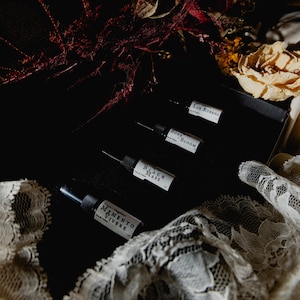 May include: A set of four small glass perfume vials arranged in a black box. Each vial features a white label with black text reading Lux Aeterna, Bone Bloom, Black Mass, and Memento Vivere. The vials have black caps and are displayed with lace.