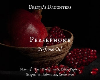 Può includere: Una ciotola di legno piena di semi di melograno rossi e un melograno intero. La ciotola è etichettata "PERSEPHONE Perfume Oil" e "FREYJA'S DAUGHTERS". Il testo sotto la ciotola recita "Notes of: Tart Pomegranate, Black Pepper, Grapefruit, Palmarosa, Cedarwood".