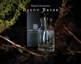 Può includere: Un flacone di profumo in vetro trasparente con tappo nero e scatola abbinata. La scatola e il flacone presentano la scritta "FREYJA'S DAUGHTERS BAYOU BRIDE". Lo sfondo è scuro con elementi naturali.