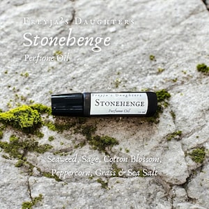 May include: A black roller bottle with a white label that reads "Freyja's Daughters Stonehenge Perfume Oil". The label also lists the scent notes: Seaweed, Sage, Cotton Blossom, Peppercorn, Grass & Sea Salt. The bottle is resting on a gray and green mossy rock.