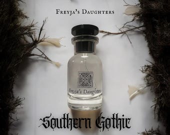 Può includere: Un flacone di profumo in vetro trasparente con tappo nero, etichettato "Freyja's Daughters" e "Southern Gothic". L'etichetta elenca le note olfattive: Gelsomino notturno, Rosa secca, Violetta ombreggiata, Radice di iris, Nag Champa e Muschio.