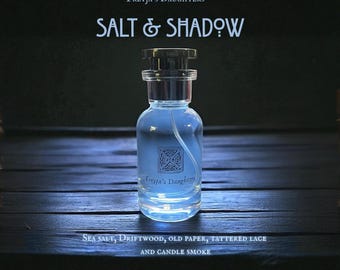 Può includere: Un flacone di profumo in vetro trasparente con tappo nero e liquido azzurro. Il flacone ha un'etichetta con un disegno a nodo celtico e il testo "Freyja's Daughters" e "SALT & SHADOW". Sul fondo del flacone c'è il testo "SEA SALT, DRIFTWOOD, OLD PAPER, TATTERED LACE AND CANDLE SMOKE".