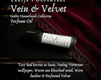 May include: A perfume oil bottle with the text "Vein & Velvet" and "Gothic Hauntlands Collection". The bottle is on a dark surface with a burgundy velvet cloth in the background. The text on the bottle reads "Freyja's Daughters".
