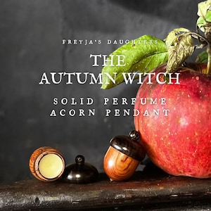 May include: A solid perfume acorn pendant with a wooden base and dark cap, shown open with yellow balm and closed next to a red apple with green leaves. Text reads: FREYJA'S DAUGHTERS THE AUTUMN WITCH SOLID PERFUME ACORN PENDANT.