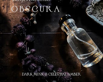 May include: A clear glass perfume bottle with a black cap, filled with a light-colored liquid. The bottle is positioned on a dark wooden surface, surrounded by dark purple flowers. The text "OBSCURA" and "DARK MUSK & CELESTIAL AMBER" are visible.