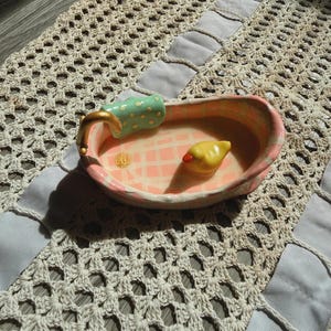 May include: A ceramic trinket dish shaped like a miniature bathtub with a pink and white checkered interior, a gold-colored faucet, a green polka-dot towel draped over the edge, and a small yellow rubber duck figurine resting inside the basin.