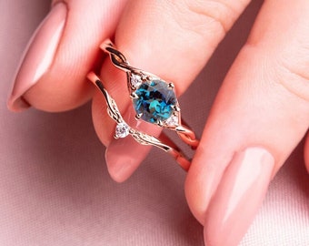 Puede incluir: Primer plano de una mano con dos anillos de oro. El anillo superior presenta una piedra preciosa azul rodeada de diamantes más pequeños. El anillo inferior es una banda delicada con un pequeño acento de diamante.