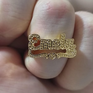 May include: A gold-colored name ring with the name "Emely" in a decorative font. The ring features a heart-shaped design between the first and last names. The ring is held by a person.
