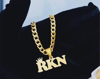 Puede incluir: Un collar dorado con una cadena de eslabones y un colgante. El colgante presenta las letras "RKN" en una fuente estilizada, con una pequeña corona encima de la "R". El collar se muestra sobre un fondo negro.