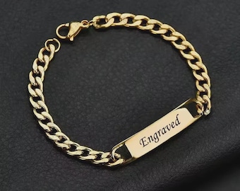 Puede incluir: Pulsera dorada con diseño de cadena de eslabones. La pulsera presenta una placa rectangular grabada con la palabra "Engraved". La pulsera se muestra sobre una superficie negra.