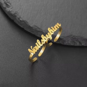 May include: A gold-colored ring with the text "Nailsbykim" in a cursive font. The ring is open and adjustable, with the text spanning across the top. The ring is displayed on a dark gray surface.