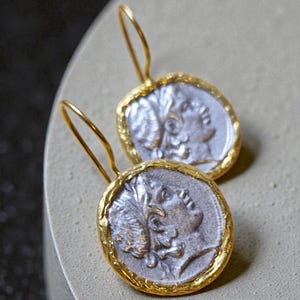 Könnte beinhalten: Ein Paar silberfarbene Münzohrringe mit goldfarbenen Rahmen und Ohrhaken. Die Münzen zeigen das Profil einer Person mit Lorbeerblättern. Die Ohrringe werden auf einer hellgrauen Oberfläche präsentiert.