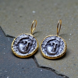 May include: A pair of silver-coloured earrings with a gold-coloured border. Each earring features a detailed face design. The earrings have a gold-coloured hook for pierced ears. The earrings are displayed on a textured, grey surface.