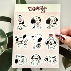 Puede incluir: Una hoja de pegatinas con perros de dibujos animados en varias poses. Los perros son principalmente blancos con acentos negros, y algunos tienen mejillas rosadas. Las pegatinas están sobre un fondo rosa cuadriculado, con la palabra "Doofys" en la parte superior.