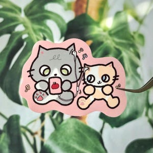 Könnte beinhalten: Ein rosa Aufkleber mit zwei Cartoon-Katzen. Eine graue Katze hört Musik mit Kopfhörern und hält ein rosa Gerät mit einem roten Apfel. Die andere Katze ist orange und weiß. Musiknoten umgeben die Katzen.