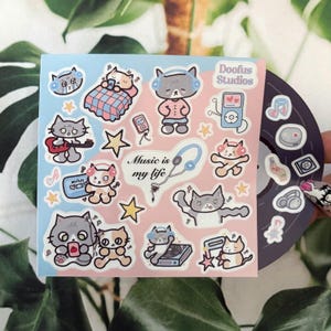 Puede incluir: Una hoja de pegatinas con temática de gatos con elementos musicales, incluyendo auriculares, notas musicales y un iPod. Las pegatinas están sobre un fondo rosa y azul, con el texto "Music is my life."