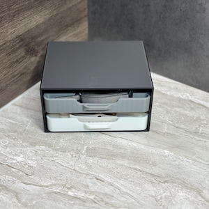 May include: A two-tiered gray and white plastic storage organizer with a gray strap in the top drawer.