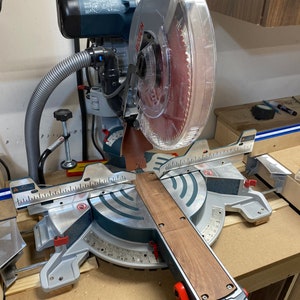 May include: A silver and black miter saw with a clear plastic guard over the blade. The saw is cutting a piece of wood. The saw has a red lever and a black handle.